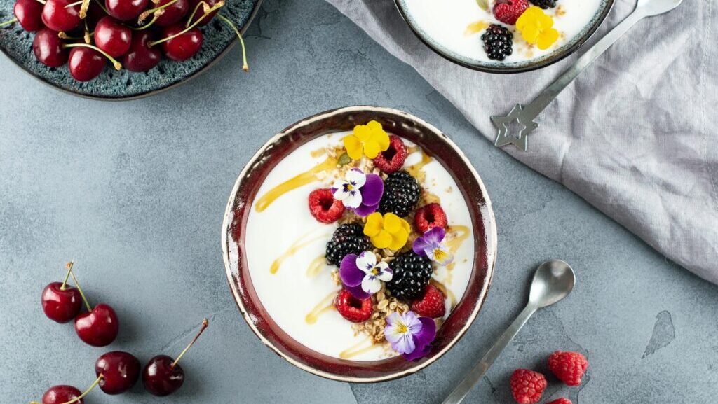 Flat lay of a vibrant yogurt bowl topped with blackberries, raspberries, and cherries, shot from above, Edible flowers for the balcony