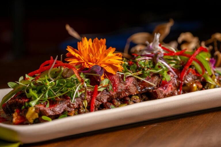 A beautifully plated gourmet beef dish garnished with vibrant edible flowers and sliced peppers.