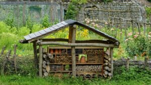hotel dla owadów, domek dla owadów DIY, A handmade insect hotel in a lush rural garden, surrounded by vibrant flowers and rustic fencing.