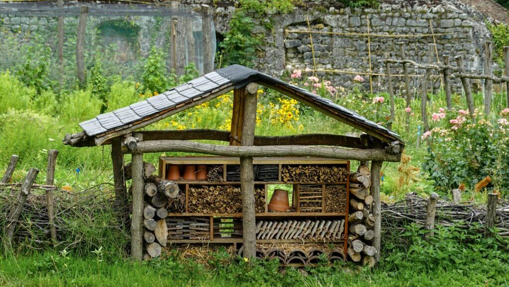 hotel dla owadów, domek dla owadów DIY, A handmade insect hotel in a lush rural garden, surrounded by vibrant flowers and rustic fencing.
