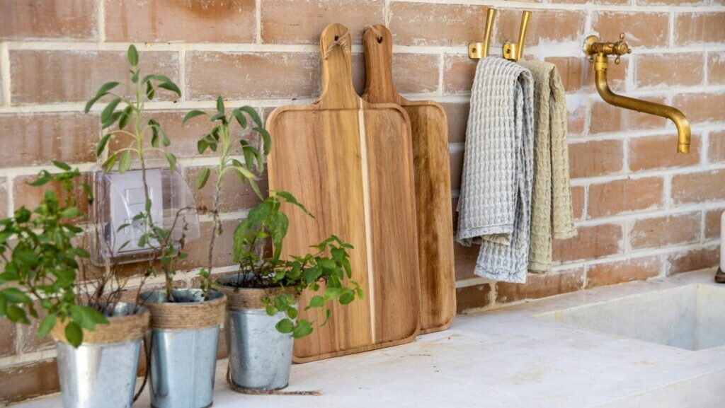kuchnia ogrodowa w stylu rustykalnym, A rustic kitchen setup featuring herb plants, cutting boards, and a brass faucet against a brick wall.